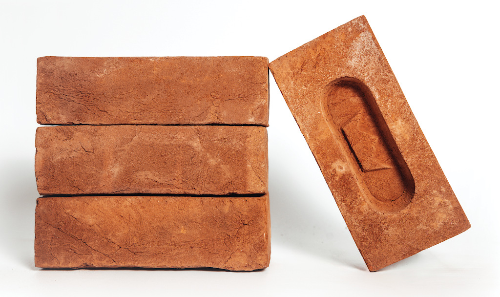 Three red handmade bricks stacked horizontally. A fourth brick leans against the right side at an angle, showing its frog indentation on the top surface.