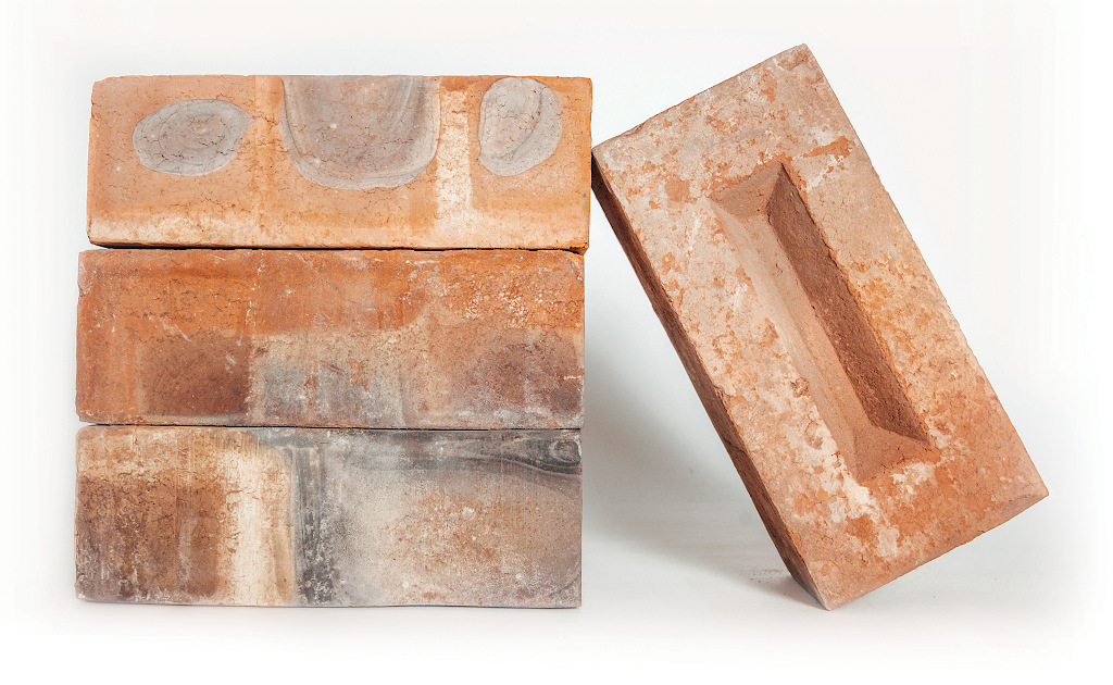 Three pressed bricks stacked horizontally. A fourth brick leans against the right side at an angle, showing its frog indentation on the top surface.