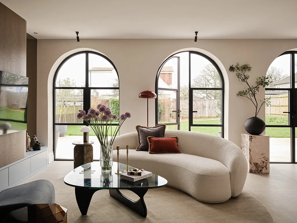 Wide view of a living room featuring a cream sofa, coffee table, and arched doorways, with one doorway open leading to the garden.