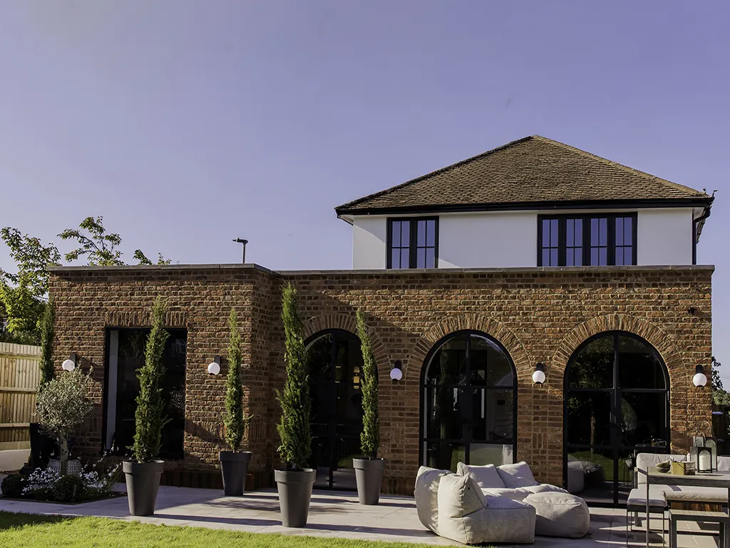 Wider view of the rear of a house from the garden, showing soft red handmade bricks, three arched doorways and outdoor patio furniture.